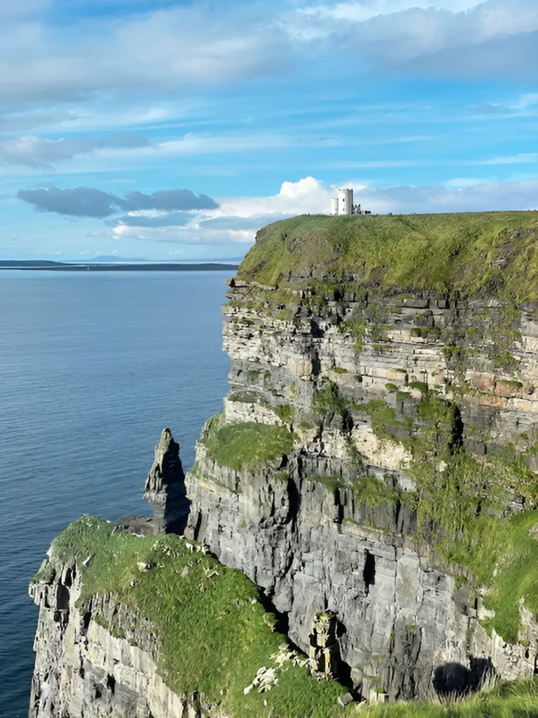 A grassy cliff with layered rocky edges rises above calm blue sea under a bright sky. A small white tower stands on top of the cliff, overlooking the ocean.